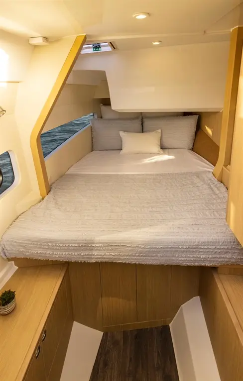 A modern boat cabin featuring a double bed with grey bedding, wooden cabinetry, and a window showing water outside.